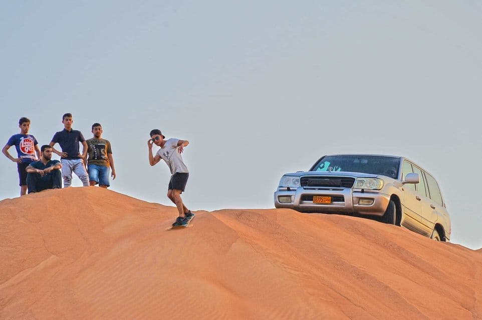 A WeRoad group trip watches as one person sandboards down a large dune, with a silver SUV parked at the top.