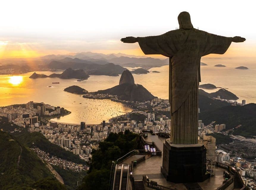 Blick von hinter einer großen Statue mit ausgebreiteten Armen, die eine weitläufige Stadt, Bucht und Berge bei Sonnenuntergang überblickt.
