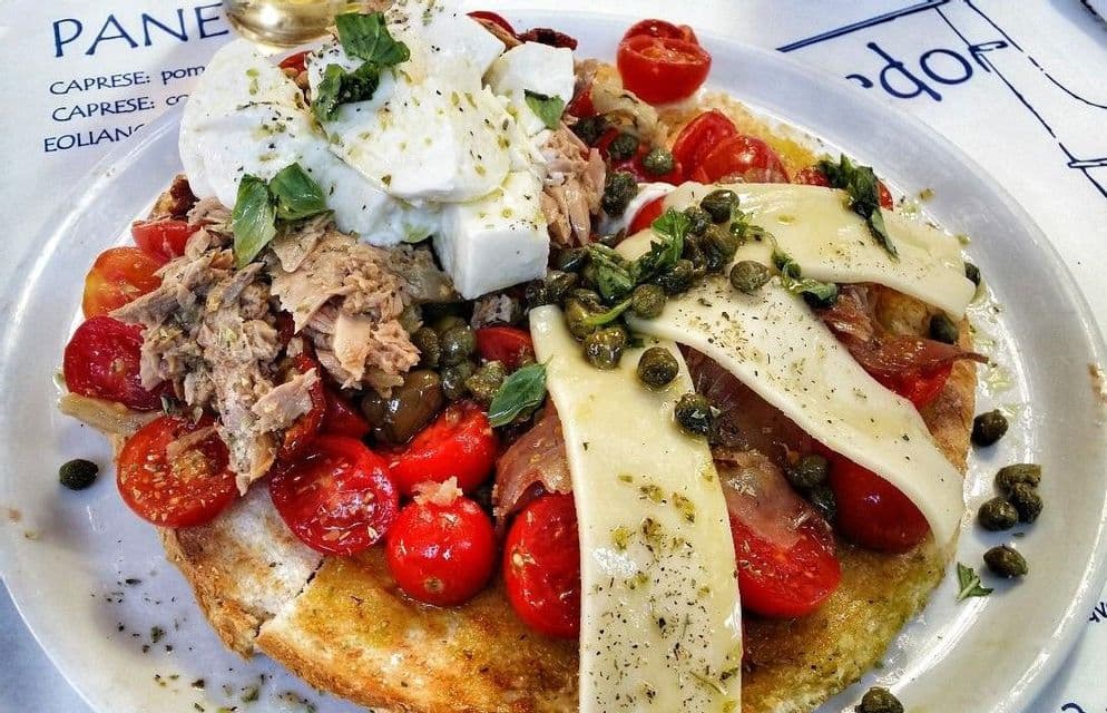 Una bruschetta grande con tonno, pomodorini, mozzarella, capperi e basilico fresco, servita su un piatto bianco.