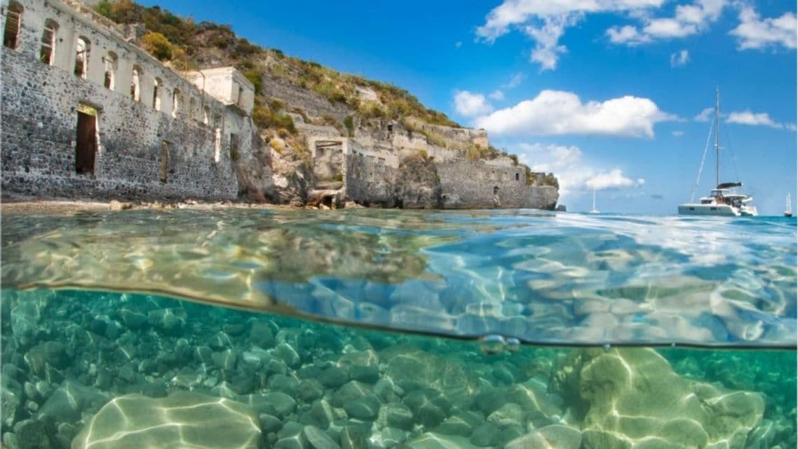 Uno scatto a filo d'acqua di un mare cristallino con fondale roccioso e rovine in pietra sulla costa, con un catamarano all'orizzonte.