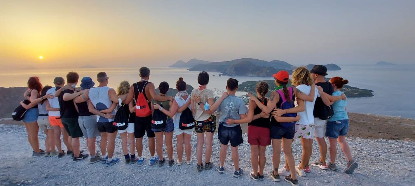 Un gruppo WeRoad in viaggio si abbraccia su un punto panoramico, ammirando il mare e le isole al tramonto.