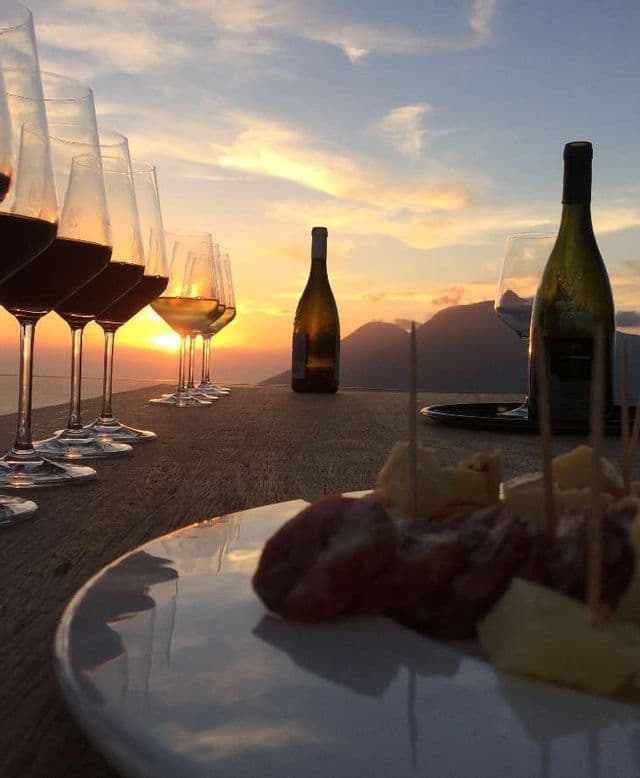 Una fila di bicchieri e bottiglie di vino su un tavolo di legno con stuzzichini, con vista sulle montagne al tramonto.