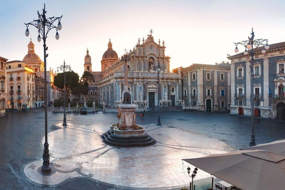 Una fuente ornamentada se alza en el centro de una amplia y vacía plaza de la ciudad frente a una gran catedral al amanecer.