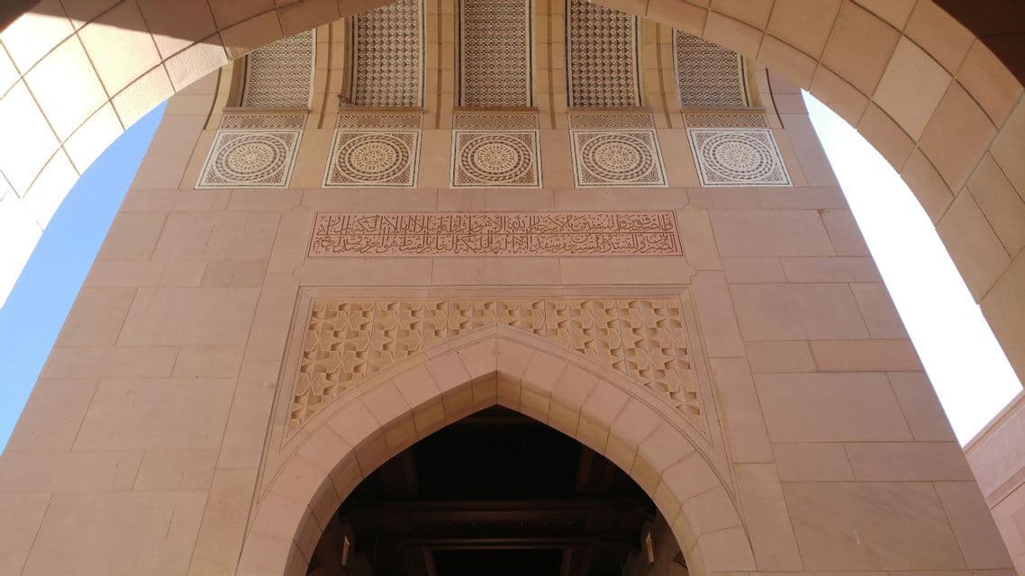 Une prise de vue en contre-plongée d'une façade de bâtiment couleur sable avec des arches complexes, des sculptures géométriques et une frise de calligraphie arabe.