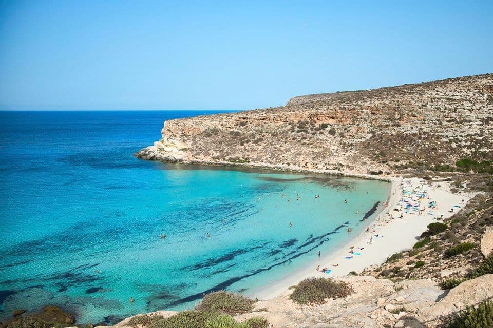 Una vista aérea de una playa de arena blanca en una cala con gente nadando en el agua clara de color turquesa, rodeada de acantilados rocosos.