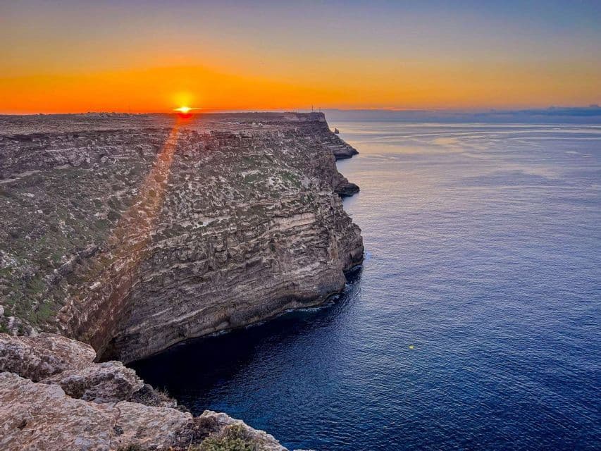 El sol se pone sobre una costa escarpada y rocosa, proyectando un cálido resplandor naranja sobre el horizonte y el profundo mar azul.