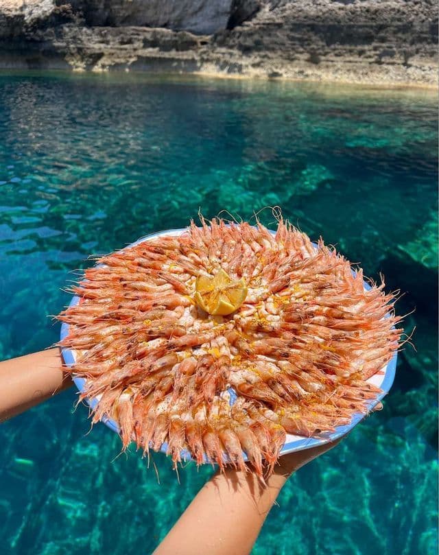 Manos sostienen una gran fuente de camarones cocidos y una rodaja de limón sobre agua turquesa cristalina, con acantilados rocosos al fondo.