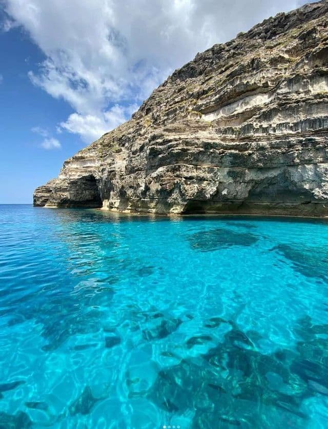 Una vista desde el agua de un acantilado rocoso estratificado que se encuentra con aguas turquesas y cristalinas bajo un cielo parcialmente nublado.