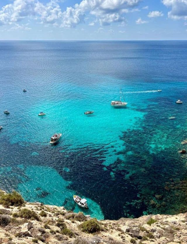 Una vista aérea de varias embarcaciones en agua turquesa y azul profundo junto a un acantilado rocoso bajo un cielo parcialmente nublado.