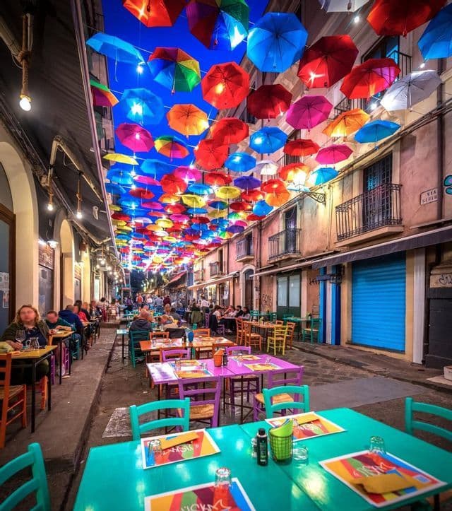 Sombrillas iluminadas y multicolores cuelgan sobre mesas de comedor al aire libre en una calle estrecha por la noche.