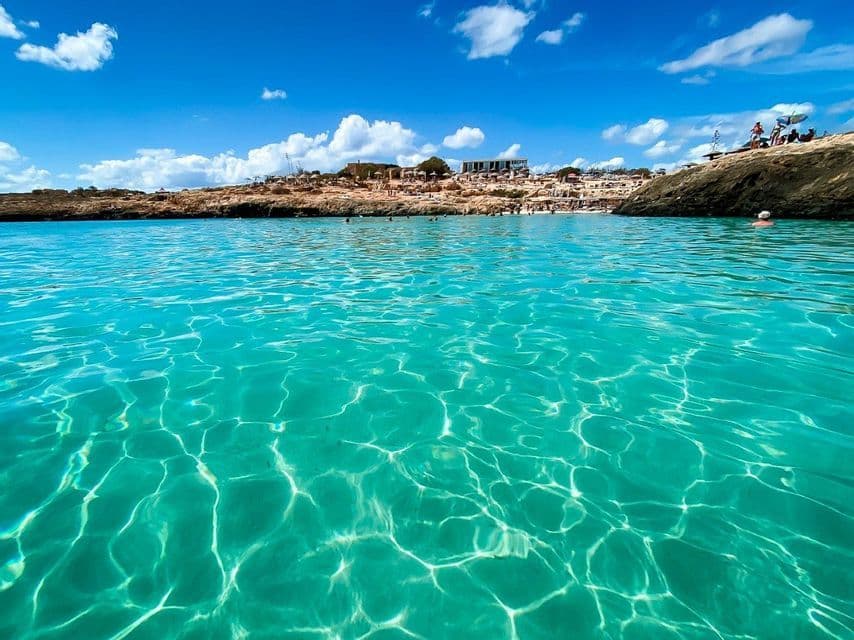 El sol se refleja en la superficie de aguas turquesas cristalinas, con una orilla rocosa abarrotada de gente al fondo.
