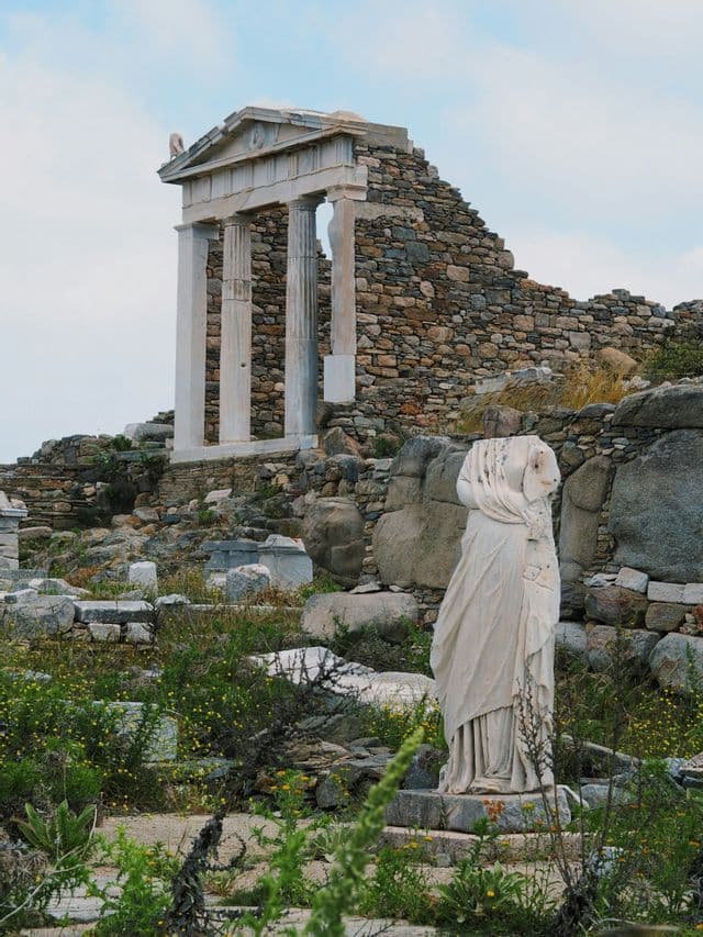 Una statua di marmo senza testa si erge tra rovine archeologiche ricoperte di vegetazione, con una facciata di tempio classico sullo sfondo.