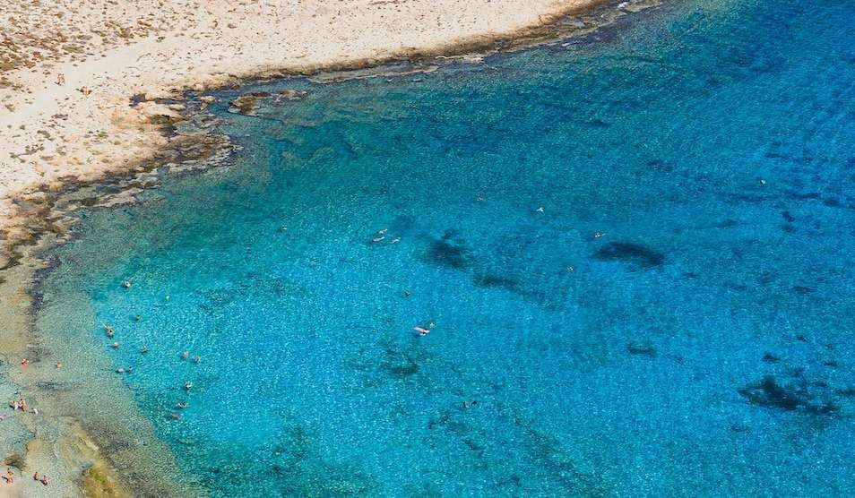 Vista aerea di una costa con spiaggia sabbiosa e persone che nuotano in acqua cristallina turchese.