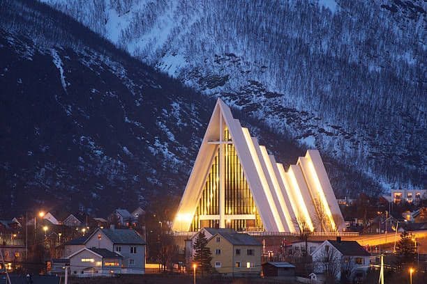 Una chiesa moderna dal design triangolare distintivo è illuminata di notte, con case residenziali sottostanti e una montagna innevata dietro.