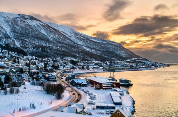 Una città costiera coperta di neve si trova ai piedi di una grande montagna, con una strada tortuosa e una baia calma al tramonto.
