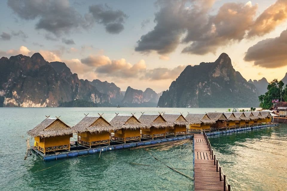 A wooden pier leads to a row of floating bungalows with thatched roofs on a calm lake, with dramatic mountains in the distance.