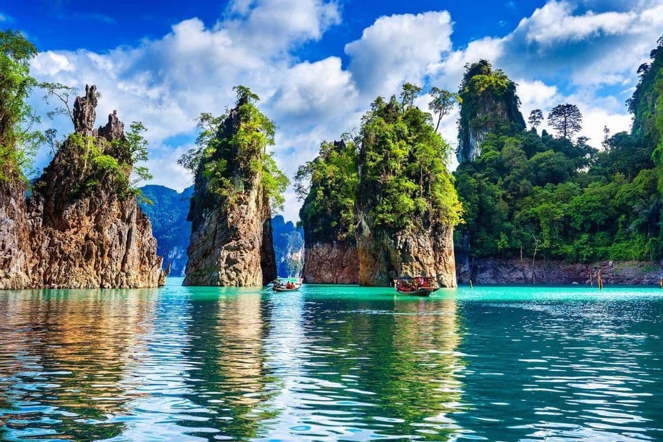 Two long-tail boats navigate turquoise waters between massive, tree-covered rock formations under a partly cloudy sky.