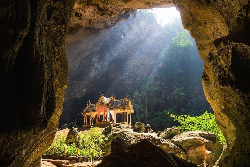A golden Thai-style pavilion sits inside a large cave, illuminated by a beam of sunlight from an opening above.
