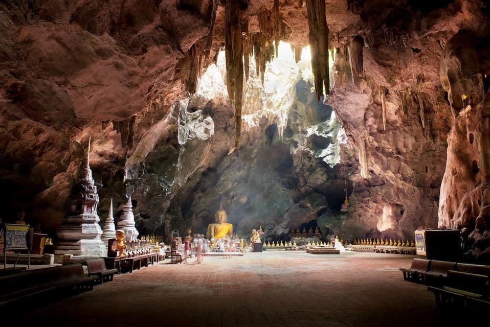 A large Buddha statue sits in a shrine inside a vast cave with stalactites, illuminated by sunlight from an opening above.