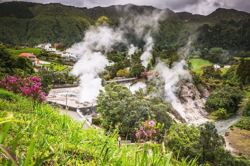 Vapore sale da sorgenti geotermiche in una rigogliosa valle verde con un villaggio e montagne ricoperte di alberi sullo sfondo.