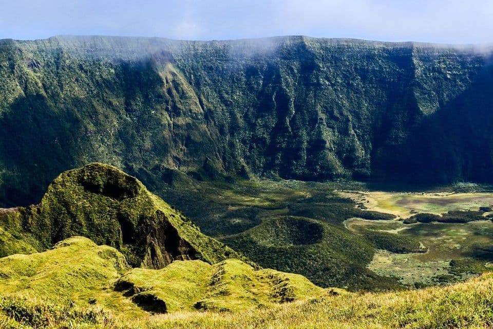 Una vista da un pendio erboso che domina una vasta e verde caldera vulcanica, con ripide scogliere boscose sotto un cielo velato.