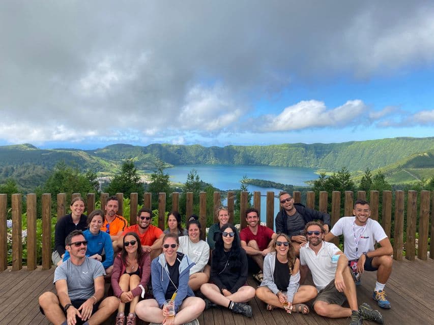 Un viaggio di gruppo WeRoad seduto insieme per una foto su una terrazza di legno con vista su un lago vulcanico e verdi colline.