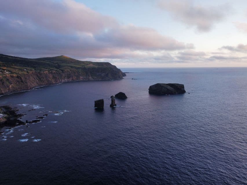 Una vista aerea di una costa frastagliata con verdi colline e grandi faraglioni che emergono dall'oceano sotto un cielo crepuscolare.