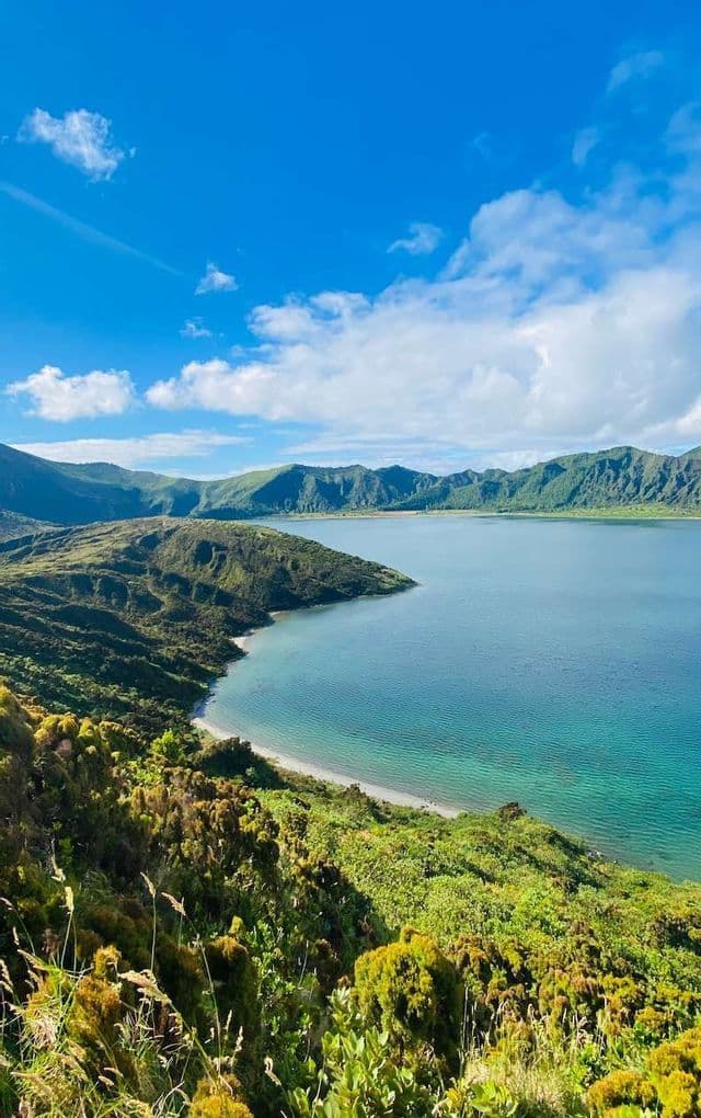 Vista dall'alto di un lago craterico turchese, circondato da lussureggianti montagne verdi, sotto un cielo azzurro e nuvoloso.