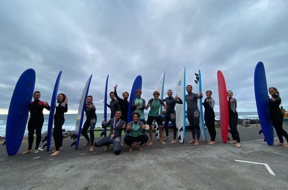 Un gruppo WeRoad in viaggio, in mute da sub, posa con le tavole da surf su un'area pavimentata vicino all'oceano sotto un cielo nuvoloso.