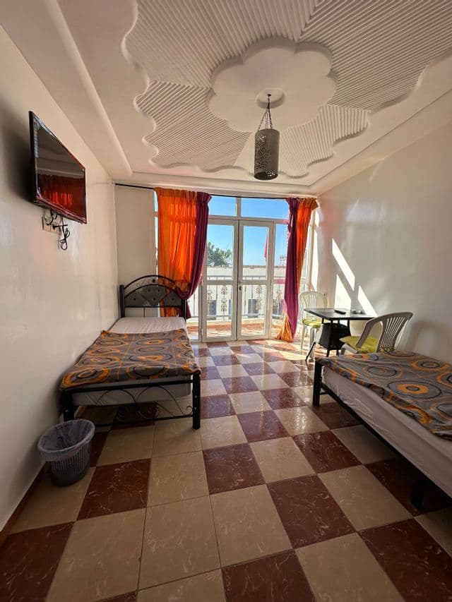 Una camera d'albergo illuminata dal sole con due letti singoli, un pavimento a scacchi e porte a vetri che si aprono su un piccolo balcone.