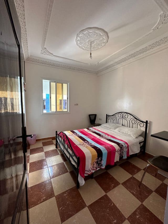 Interno di una camera semplice con letto in ferro battuto, coperta a righe colorate e pavimento in piastrelle a scacchi marroni e beige.