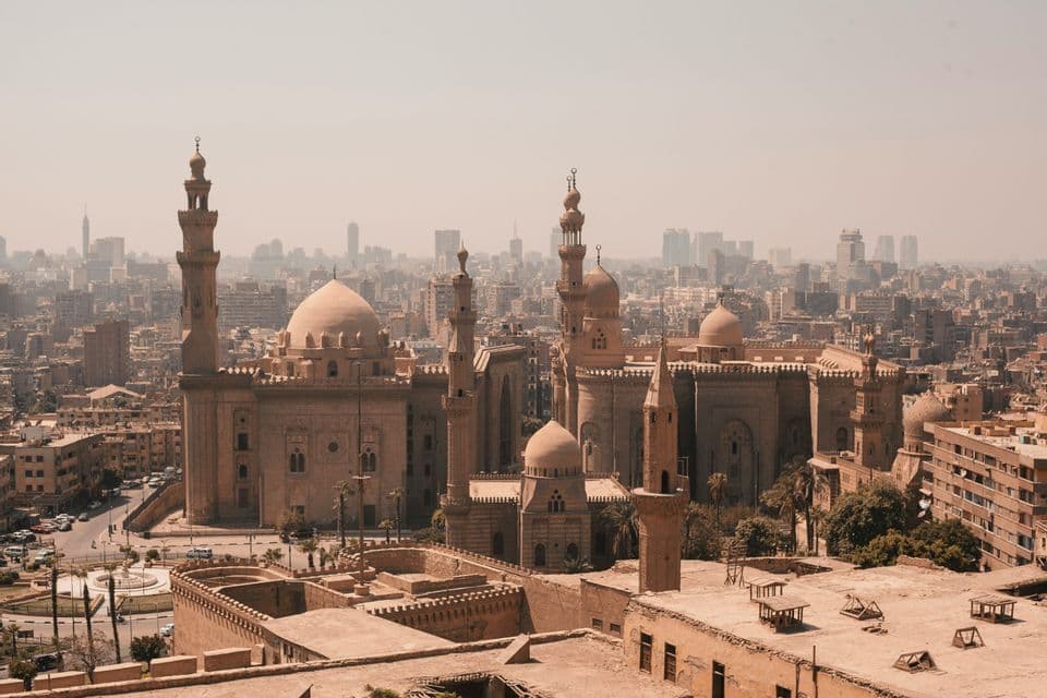 Un esteso complesso di moschee con minareti e cupole, visto dall'alto sullo sfondo di uno skyline moderno e leggermente sfocato.