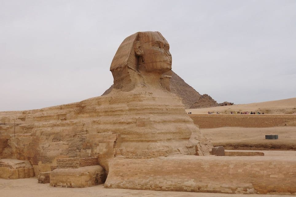 La statua della Grande Sfinge è vista di lato, con una grande piramide visibile sullo sfondo nel deserto sotto un cielo nuvoloso.