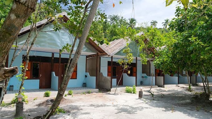 Una hilera de bungalows azul claro con puertas de madera y pequeños porches, ubicados entre exuberantes árboles verdes en terreno arenoso.