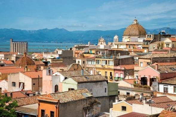 Un panorama urbano di edifici colorati con tetti in terracotta, una grande cupola, il mare e montagne distanti sotto un cielo azzurro.