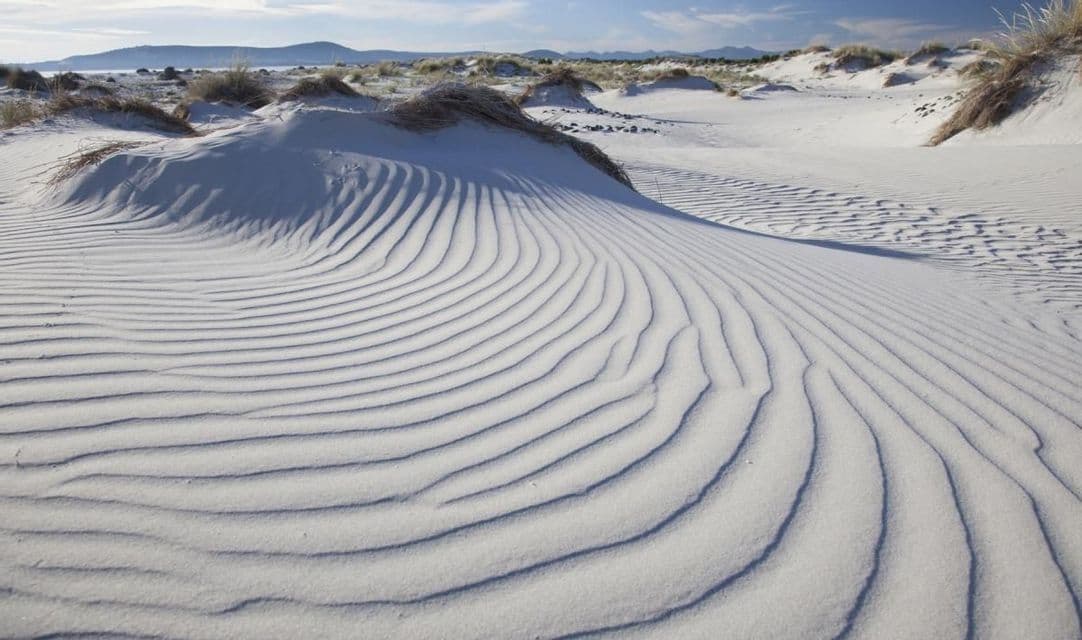 Ondulazioni create dal vento sulle dune di sabbia bianca, con colline visibili in lontananza sotto un cielo azzurro.