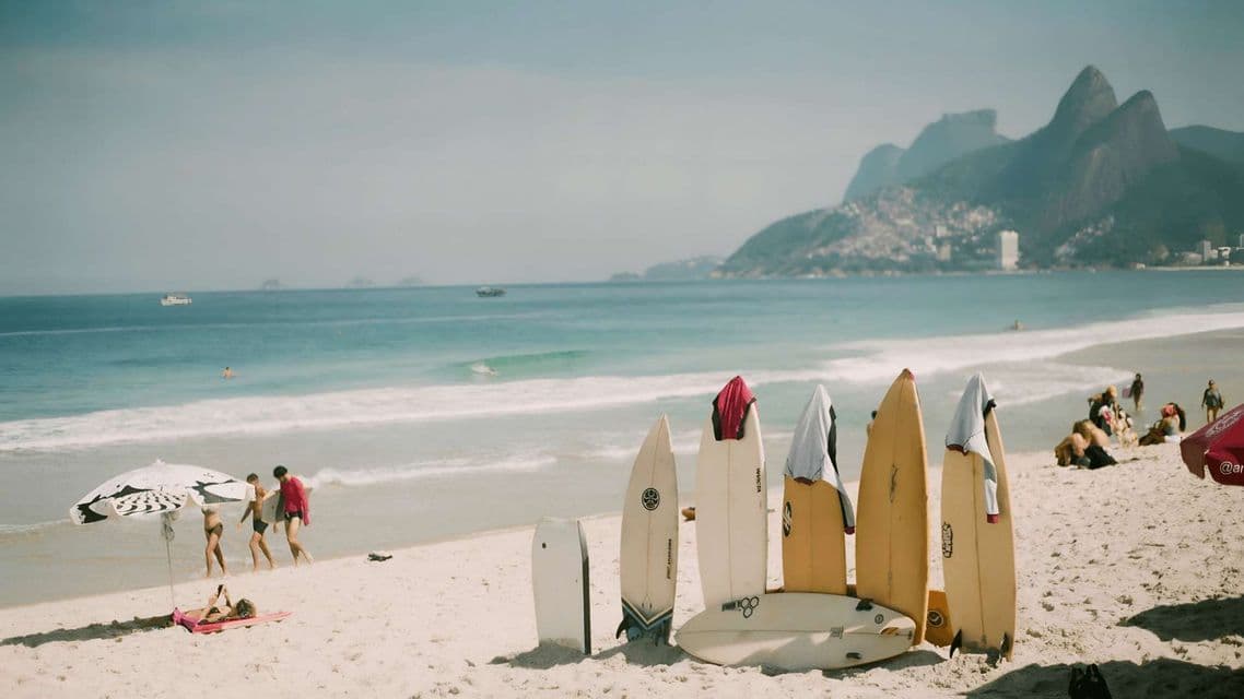 Varias tablas de surf están de pie en la arena de una playa soleada, con gente relajándose junto al océano y montañas al fondo.