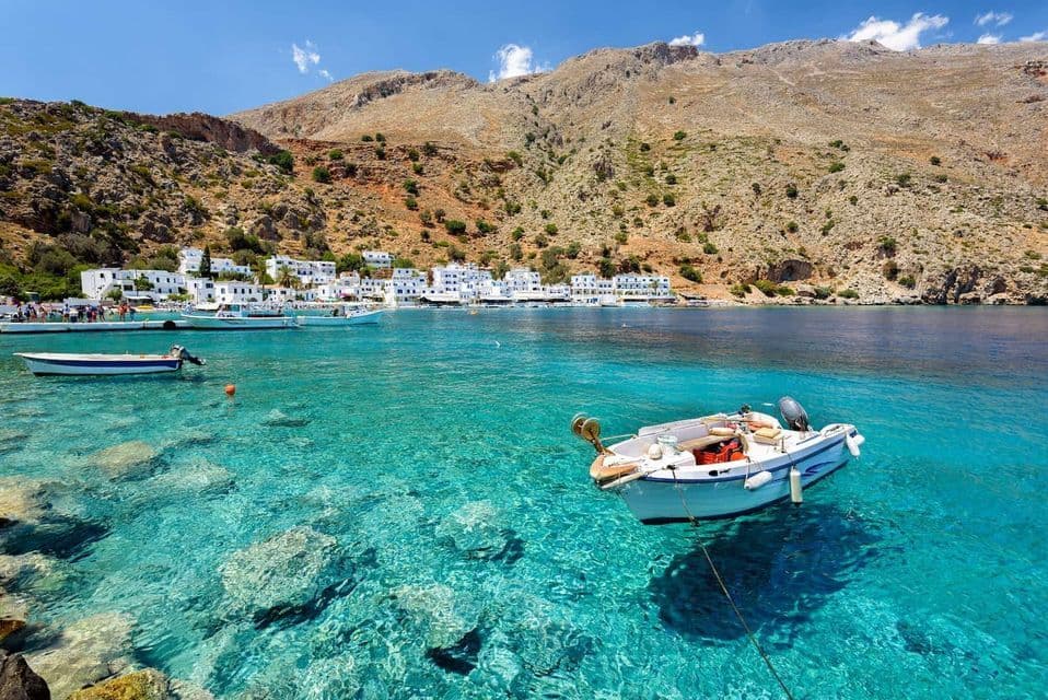 A small boat is anchored in clear, turquoise water in front of a coastal village with white houses at the base of a rocky mountain.