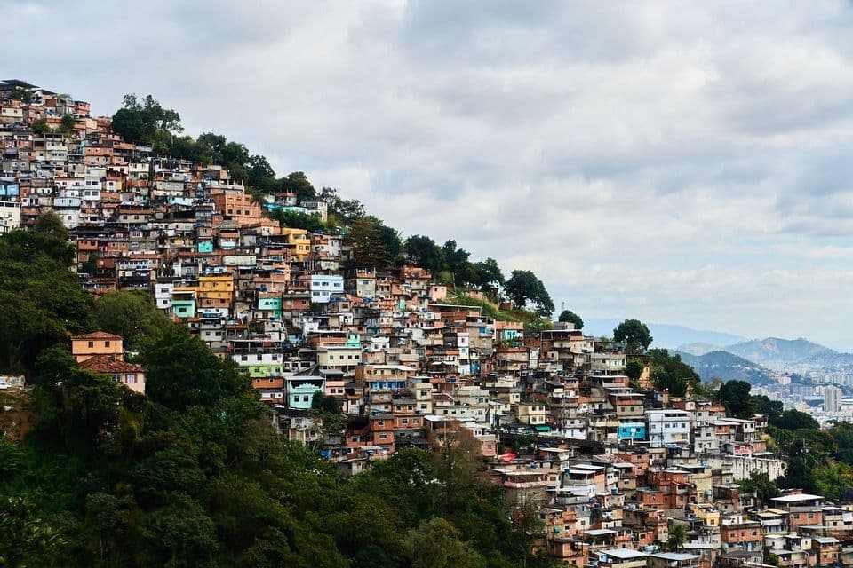 Des maisons colorées et serrées recouvrent un flanc de colline escarpé et verdoyant sous un ciel nuageux, avec des montagnes visibles au loin.