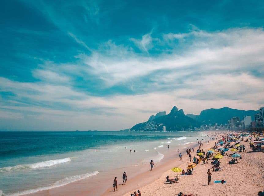 Ein belebter Sandstrand, an dem Menschen unter bunten Sonnenschirmen schwimmen und entspannen, mit Bergen und einer Stadt im Hintergrund.