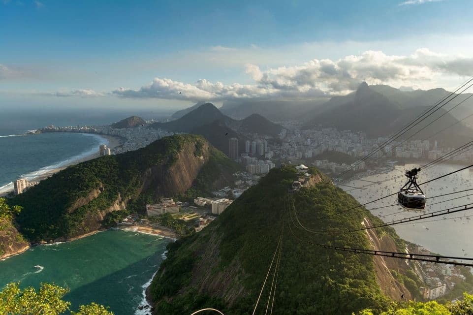 Un teleférico recorre cables en lo alto sobre una ciudad costera, enclavada entre exuberantes montañas verdes y el océano.