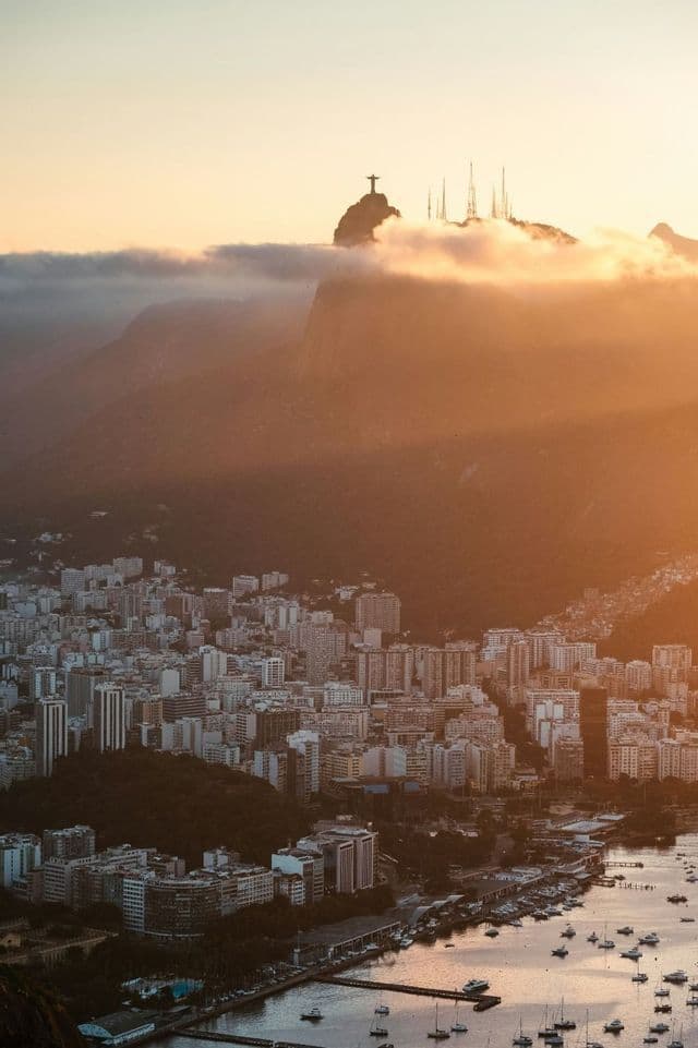 Eine ausgedehnte Küstenstadt und eine Bucht mit Booten bei Sonnenaufgang, mit der Silhouette einer großen Statue auf einem fernen Berggipfel.