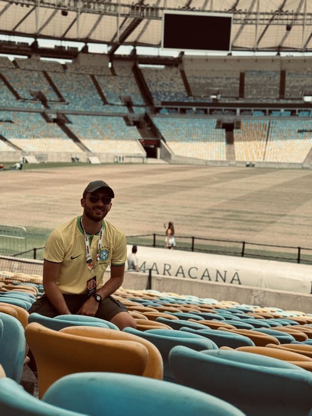 Un homme souriant, vêtu d'un maillot jaune et d'une casquette, est assis dans les tribunes vides d'un grand stade.