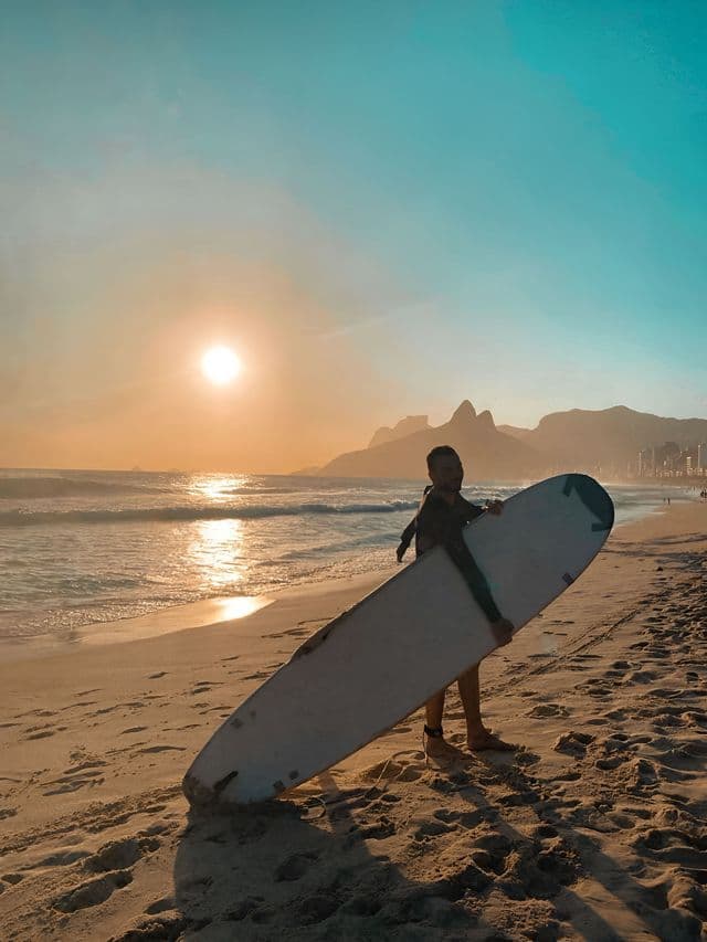 Ein Surfer im Neoprenanzug hält ein Surfbrett an einem Sandstrand, während die Sonne über dem Meer und fernen Bergen untergeht.