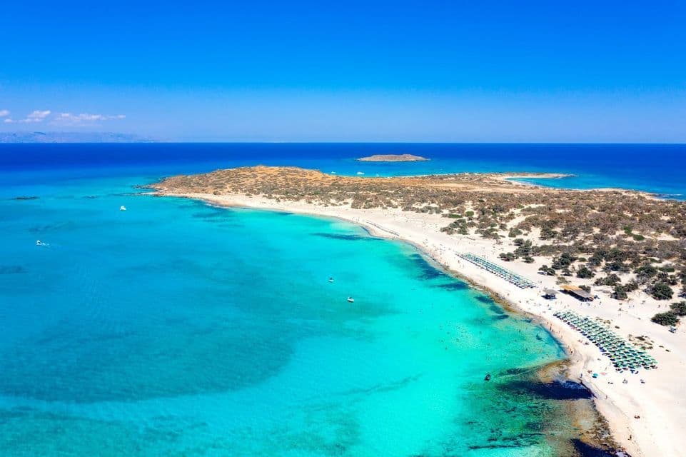 Vue aérienne d'un banc de sable reliant une petite île, avec une plage de sable blanc bordée de parasols et des eaux turquoise claires.