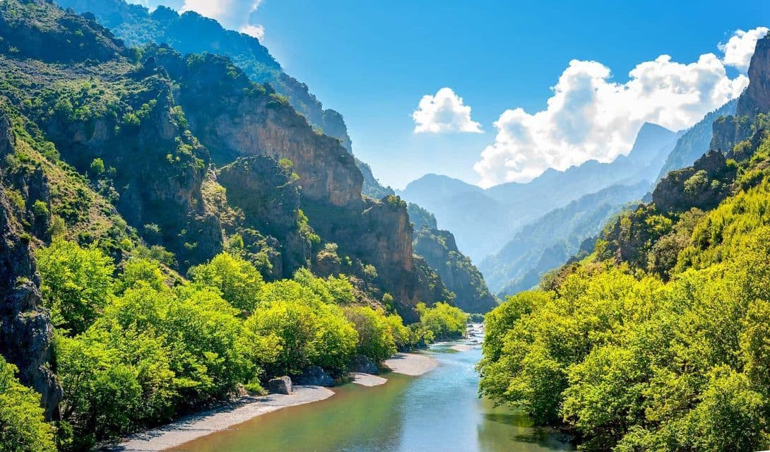 Un río serpentea por un exuberante valle de montaña verde con acantilados escarpados, bajo un cielo azul y nubes blancas.