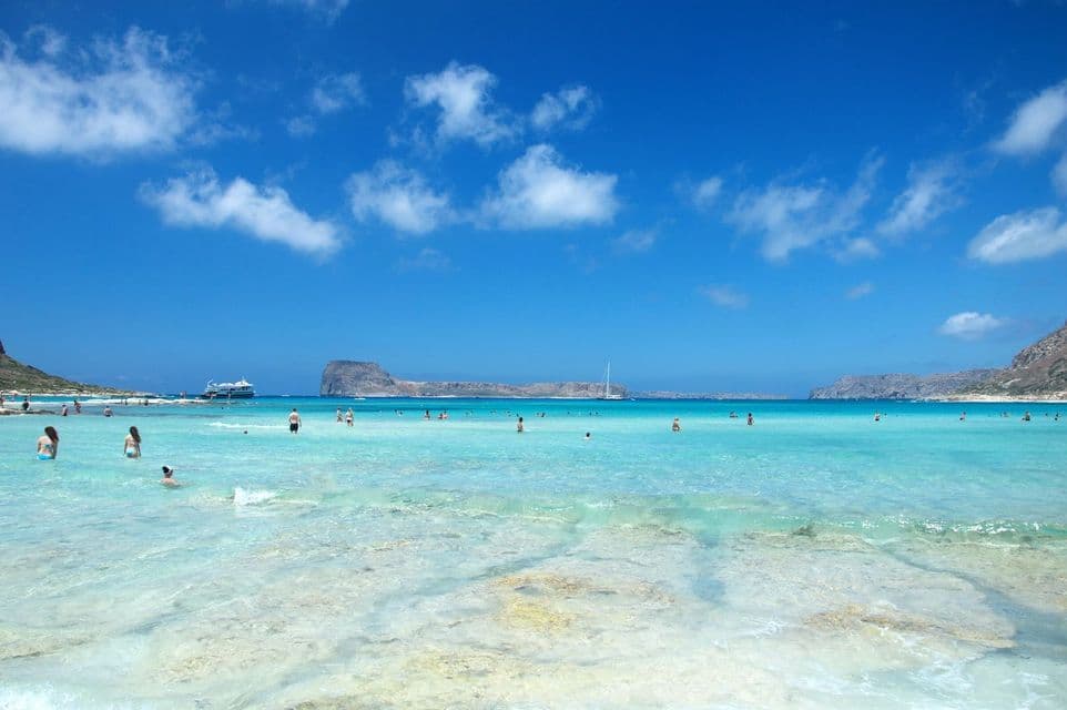 People swimming and relaxing in shallow, clear turquoise water at a beach, with rocky islands under a blue sky with white clouds.