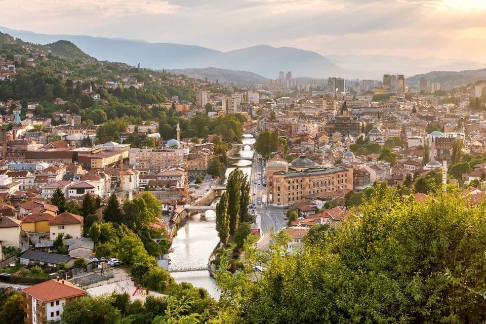 Un paesaggio urbano visto dall'alto al tramonto, con un fiume e diversi ponti che lo attraversano al centro e le montagne sullo sfondo.
