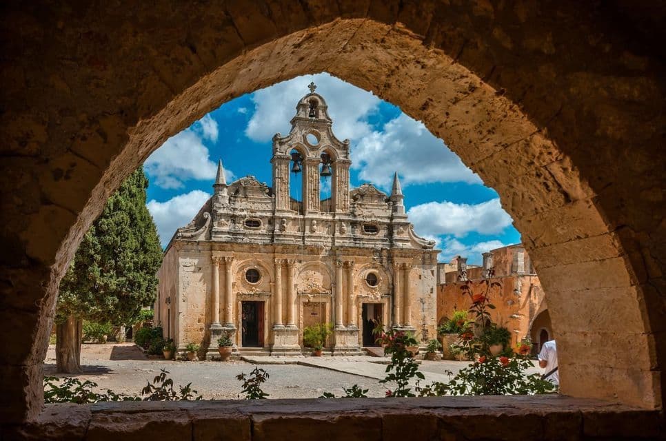 Una iglesia de piedra ornamentada con un campanario, vista desde el interior de un oscuro arco de piedra en un día soleado.