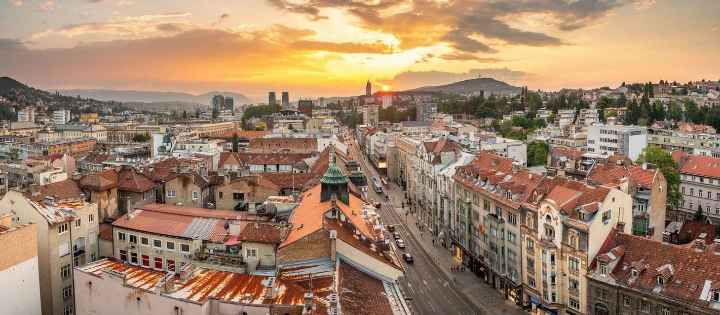 Una vista elevada y panorámica de un extenso paisaje urbano con tejados rojos bajo un cielo de atardecer dorado.